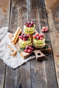 Drei Emmentaler-Käse-Barrels mit Trauben und Kräutern, serviert mit Brotstangen auf einem rustikalen Holzbrett.