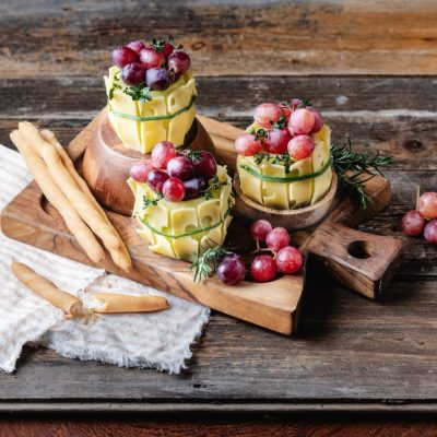 Drei Emmentaler AOP Käse Weinfässer mit Trauben und Kräutern, mit Grissinistangen auf einem rustikalen Holzbrett.