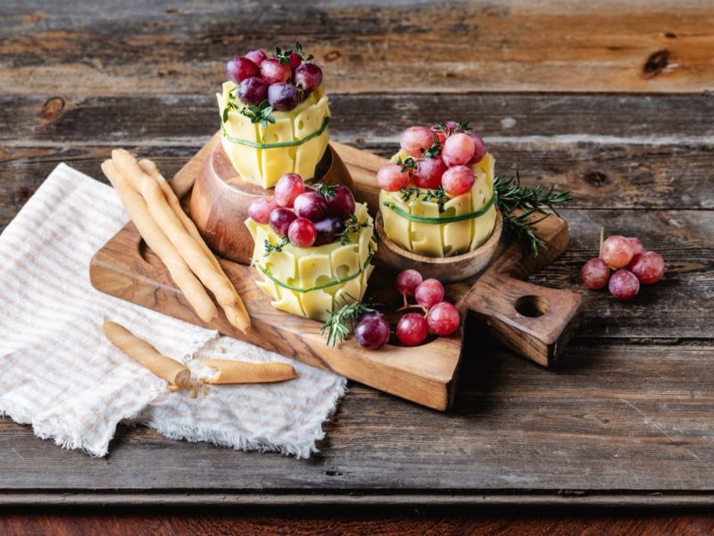 Drei Emmentaler AOP Käse Weinfässer mit Trauben und Kräutern, mit Grissinistangen auf einem rustikalen Holzbrett.